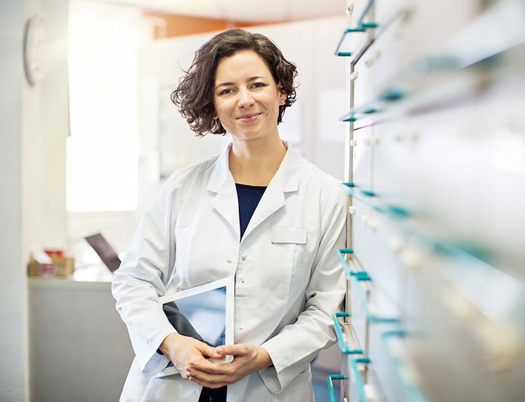 Portrait of confident young female pharmacist leaning to a medicine shelf with a digital tablet